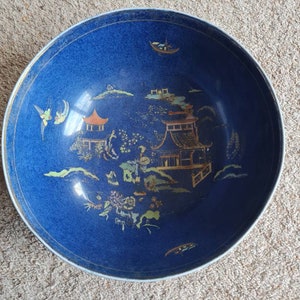 Könnte beinhalten: Eine große blaue Keramikschüssel mit weißem Rand und handbemaltem Design einer chinesischen Landschaft mit goldenen Akzenten. Das Design umfasst eine Pagode, Bäume und Vögel.