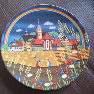 Può includere: Un piatto decorativo rotondo con una colorata illustrazione di un villaggio con una chiesa, case e un arcobaleno nel cielo. Il primo piano mostra un campo di grano con due figure intente al raccolto, circondate da spighe, margherite e fiori rossi.