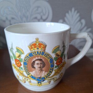 Op de afbeelding: Een witte keramische mok met een kleurrijk beeld van koningin Elizabeth II en het koninklijke wapen. Het wapen bevat de woorden "Dieu et mon droit" en "Honi soit qui mal y pense".