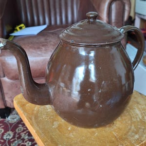 以下が含まれることがあります： 蓋と取っ手が付いた、茶色のヴィンテージスタイルのティーポット。 ティーポットは金属製で、木製の表面に置かれています。 丸みを帯びたボディと湾曲した注ぎ口があります。