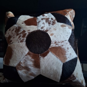 May include: A square-shaped pillow with a patchwork design made of different colored cowhide. The pillow is made of brown, white, and black cowhide. The pillow has a star-shaped design in the center.