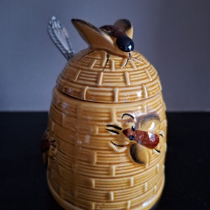 May include: A ceramic honey pot in the shape of a beehive, with a bee figurine on the lid. The pot is tan with a woven texture, and a silver spoon is placed in the lid. Decorative bees are painted on the side.
