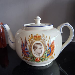 May include: A cream-coloured ceramic teapot featuring a central portrait of a man in profile, surrounded by a crown, flags, and floral motifs. The text "LONG MAY HE REIGN" is printed around the portrait. The teapot has blue trim on the lid and handle.