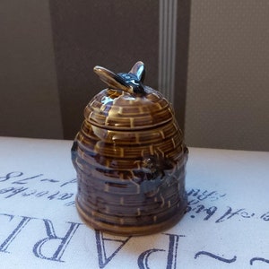 May include: A brown ceramic honey pot with a lid, shaped like a beehive. The pot has a textured, woven design and a small bee figurine on top. The pot is glazed with a glossy finish.