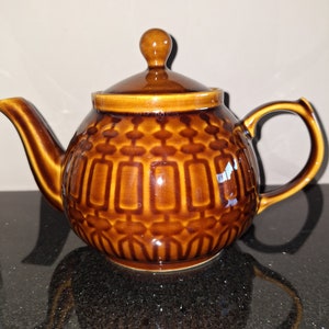 May include: A brown ceramic teapot with a geometric pattern. The teapot has a lid with a knob on top and a spout on one side. The teapot is sitting on a black surface.
