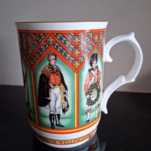 May include: White ceramic mug featuring illustrations of historical figures in military attire. The mug has a decorative rim with orange and green patterns. The handle is white and has a unique, curved design. The words "DUKE OF WELLINGTON" are printed on the mug.