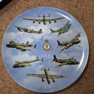 Pode incluir: Um prato de cerâmica azul e branco com uma borda dourada apresenta uma coleção de oito aeronaves militares vintage, incluindo um bombardeiro, um caça e um avião de reconhecimento. As aeronaves são pintadas em cores de camuflagem e voam em formação sobre um fundo de céu azul. O prato também apresenta o brasão da Royal Air Force.
