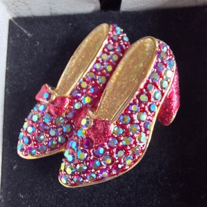 Peut inclure: Une paire de broches en forme de pantoufles rouges ornées de bijoux avec des accents dorés. Les broches sont recouvertes de strass rouges et verts irisés.