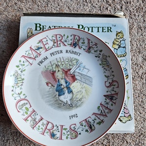 May include: A white decorative plate with a red rim, featuring Peter Rabbit and the text "MERRY CHRISTMAS from PETER RABBIT 1992." The plate is decorated with illustrations of rabbits and foliage. A book with the words "BEATRIX POTTER" is visible in the background.