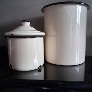 Puede incluir: Dos botes de almacenamiento vintage de esmalte color crema con ribetes negros. Uno tiene tapa, el otro es más alto y cilíndrico. Los botes están sobre una superficie negra, reflejando la luz.