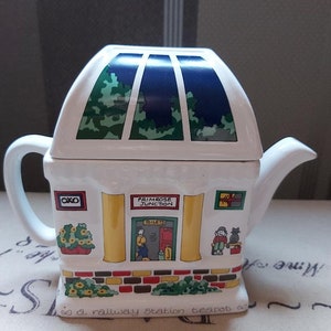 Może przedstawiać: Biały ceramiczny czajnik w kształcie dworca kolejowego z zielonym i niebieskim dachem. Czajnik ma tabliczkę z napisem "Primrose Junction" i małą tabliczkę z napisem "OXO".