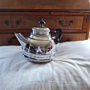 Può includere: Una teiera d'argento con un manico nero. La teiera ha una superficie lucida e riflettente e un corpo rotondo. La teiera è appoggiata su una superficie bianca.