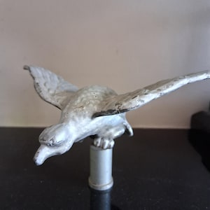 Op de afbeelding: Een zilverkleurig metalen vogelbeeldje met wijd gespreide vleugels in vluchtpositie, gemonteerd op een cilindrische voet. Het oppervlak van de vogel vertoont tekenen van slijtage en textuur, met een naar beneden gerichte kop en gedetailleerde veren.