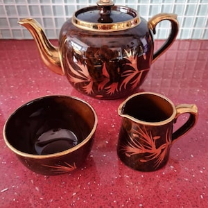 Puede incluir: Un juego de té marrón vintage con detalles dorados. El juego incluye una tetera, un azucarero y una jarrita para la leche. La tetera tiene tapa y un pico curvo. El juego está decorado con un diseño floral dorado.