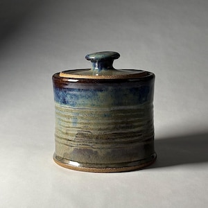 May include: A cylindrical ceramic jar with a matching lid featuring a knob handle. The exterior displays a reactive glaze with horizontal bands of deep blue, muted green, and earthy brown tones, showcasing a smooth, glossy finish throughout.