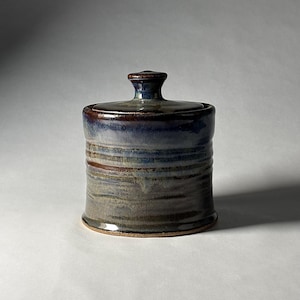 May include: A cylindrical ceramic jar with a matching lid featuring a knob handle. The exterior has a glossy glaze with blended tones of deep blue, earthy brown, and muted grey, showcasing horizontal ridges along the body of the vessel.