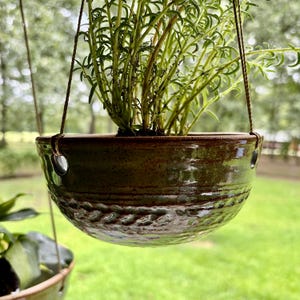 May include: A round, glazed ceramic hanging planter with a deep brown and green mottled finish. The bowl features a textured, indented band around the lower half and two side holes for suspension cords, containing a vibrant green leafy plant.