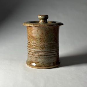 May include: A cylindrical ceramic jar with a lid, featuring a speckled brown and muted green glaze. The body of the jar has horizontal ribbed textures around the center, a wide flat rim, and a small knob handle on the center of the lid.