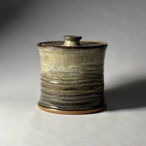 May include: A cylindrical ceramic jar with a lid, featuring a gradient glaze that transitions from a light cream color at the top to dark brown and earthy tones at the base, with visible horizontal ridges along the body of the vessel.