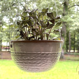 May include: A hanging ceramic planter with a speckled light gray glaze and a textured horizontal ribbed pattern around the lower half. The bowl-shaped vessel features a smooth brown rim and is suspended by thin white cords containing a green plant.