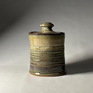 May include: A cylindrical ceramic jar with a matching lid, featuring a variegated glaze in shades of olive green, tan, and dark brown. The exterior surface displays horizontal ridged textures and a smooth, glossy finish throughout.
