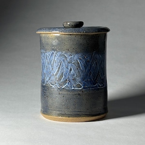May include: A handmade ceramic jar with a lid. The jar is cylindrical with a speckled gray and blue glaze. A band of blue and white wavy lines encircles the middle. The lid has a small knob and the base is unglazed.