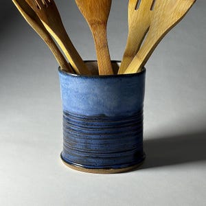 May include: A blue ceramic utensil holder with a textured, horizontal stripe design. The holder contains several wooden cooking utensils. The color of the holder transitions from a lighter blue at the top to a darker blue at the bottom.