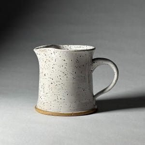 May include: A speckled white ceramic pitcher with a handle. The pitcher has a wide mouth and a slightly flared base. The exterior is covered in small, dark brown speckles. The base is a light tan color.