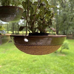 May include: A hanging ceramic planter with a two-tone finish, featuring a smooth, dark olive-green glazed upper section and a textured, tan-colored lower half. The bowl is suspended by thin cords attached to small, circular metal hardware.