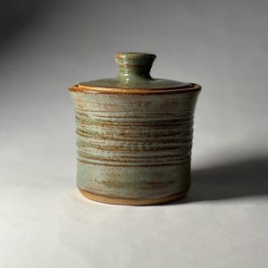 May include: A cylindrical ceramic jar with a lid, featuring a light sage green glaze with horizontal brown textured bands and speckles. The container has a flat base and a small knob handle on the center of the lid, showcasing a handcrafted finish.
