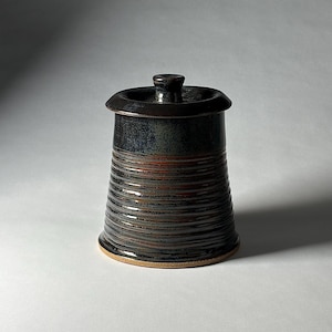 May include: A handcrafted ceramic jar with a lid. The jar has a dark brown, textured exterior with horizontal ridges and a contrasting lighter brown base. The lid is a darker shade of brown with a small knob.