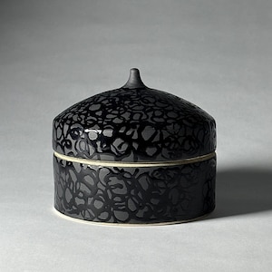 May include: A black ceramic lidded jar with a textured, floral-like pattern. The jar has a rounded shape with a small knob on the lid. The base is a lighter color, creating a contrast with the dark body.