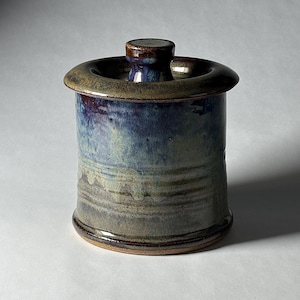 May include: A cylindrical ceramic jar with a lid, featuring a glossy glaze in shades of deep blue, muted green, and brown. The vessel has a wide, flat rim and a small, knob-like handle on the center of the lid, with visible horizontal texture.