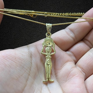 May include: Gold-toned pendant necklace featuring a stylized figure holding a circular object above their head. The pendant is suspended from a delicate gold chain. The figure has a slender form and is standing upright. The necklace is displayed against a dark background.