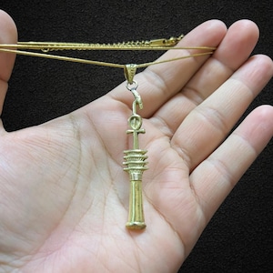 Op de afbeelding: Een goudkleurige hangerketting met een Ankh-symbool en een gestileerd kolomontwerp. De hanger hangt aan een delicate gouden ketting. De ketting wordt tegen een donkere achtergrond gehouden.