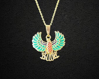 Puede incluir: Collar dorado con un colgante que representa un pájaro estilizado y colorido con las alas extendidas. Las alas del pájaro son de color turquesa, con detalles en rojo y azul. El collar cuelga sobre un fondo negro.