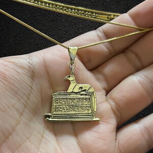 Może przedstawiać: Złoty naszyjnik z wisiorkiem w stylu egipskim. Wisiorek przedstawia stylizowaną postać ptaka na prostokątnej podstawie z hieroglifami. Łańcuszek naszyjnika jest cienki, w kolorze złotym.