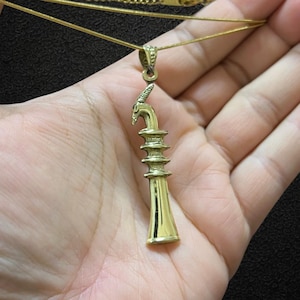 Op de afbeelding: Een goudkleurige hangerketting met een gedetailleerd, gestileerd ontwerp. De hanger heeft een gelaagde structuur met een gebogen bovenelement. De ketting is een fijne, dunne gouden kleur. De achtergrond is een donkere, effen kleur.