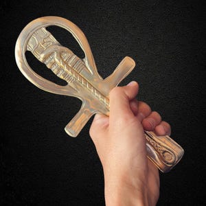 Può includere: Un simbolo Ankh color oro, un antico simbolo geroglifico egizio della vita, è tenuto su uno sfondo nero. L'Ankh presenta un anello in alto, una linea verticale e braccia orizzontali. Il simbolo è dettagliato con motivi scolpiti.