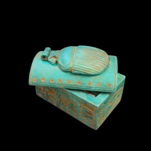 Può includere: Una scatola decorativa turchese con coperchio a cerniera, con un design a scarabeo. La scatola è adornata con intagli geroglifici e ha una forma rettangolare. Lo sfondo è nero.