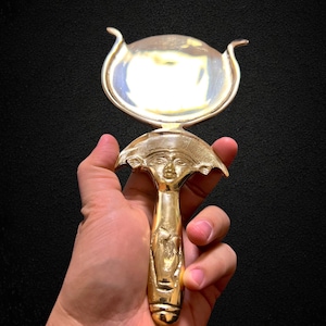 Puede incluir: Un espejo de mano dorado y ornamentado, con un mango detallado y una superficie redonda y reflectante. El mango presenta una cara esculpida y otros diseños intrincados. El espejo se sostiene sobre un fondo oscuro.
