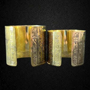 Puede incluir: Dos pulseras de puño de metal dorado. Las pulseras tienen un diseño abierto y están grabadas con símbolos jeroglíficos. Las pulseras se muestran sobre un fondo negro.