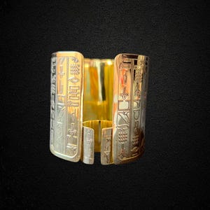 May include: A gold-colored cuff bracelet with engraved hieroglyphs. The bracelet has a rectangular shape with a split design, featuring a polished gold interior and a detailed exterior with ancient Egyptian symbols. The background is black.