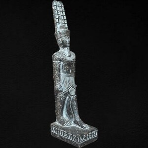 Puede incluir: Estatua de piedra gris oscuro de un faraón egipcio. La estatua está de pie con una pierna hacia adelante y lleva una alta y ornamentada tocado. La figura está detallada con líneas y patrones tallados, y se encuentra sobre una base rectangular.