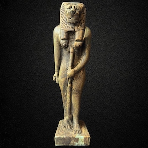 Może przedstawiać: Szczegółowa statua egipskiego bóstwa z głową lwa i ludzkim ciałem. Postać trzyma laskę i stoi na prostokątnej podstawie. Statua ma zwietrzały, postarzały wygląd z zielonkawym odcieniem.