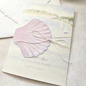 May include: A wedding invitation featuring a watercolor scene of a coastal town with sailboats. A light purple seashell-shaped embellishment is tied with string. The invitation text includes the words "Bristol, Rhode Island" and "reception to follow."