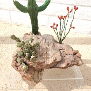 May include: A handcrafted desert scene featuring a green wire cactus with outstretched arms, a cluster of red-orange flowers, and a small plant with white flowers. The plants are set on a textured rock formation, displayed on a clear acrylic stand.