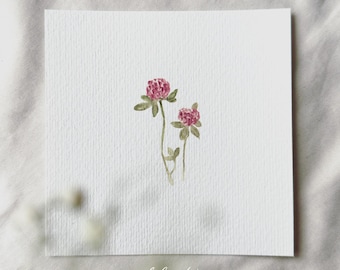 May include: A watercolor painting of two pink clover flowers with green stems and leaves on a white background. The painting is signed "delicate aesthetic" in the bottom right corner.