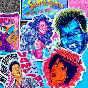 May include: A collection of colorful stickers featuring pop art portraits and text. The stickers include phrases like "Santa Carla, Murder Capital of the World," "Cry Little Sister," and "Maggots, You're Eating Maggots." The art style is vibrant and graphic.
