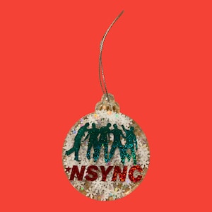 May include: Clear Christmas bauble with a silver string, featuring a green silhouette of five people and the word "NSYNC" in red glitter. The bauble is filled with white glitter and snowflakes, set against a red background.